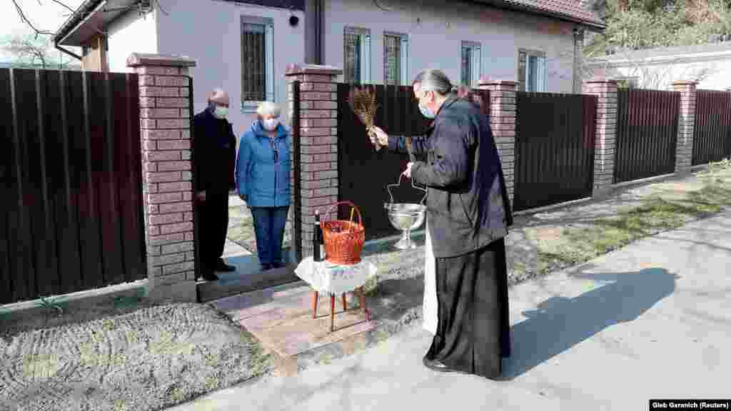 Священник ПЦУ в селе под Киевом благословляет пасхальные куличи и яйца у двора пожилой пары. Украина, 19 апреля